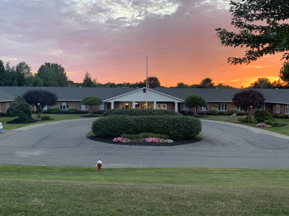 Front entrance of a single-story senior living building with a circular driveway, landscaped bushes and a flagpole at sunset.