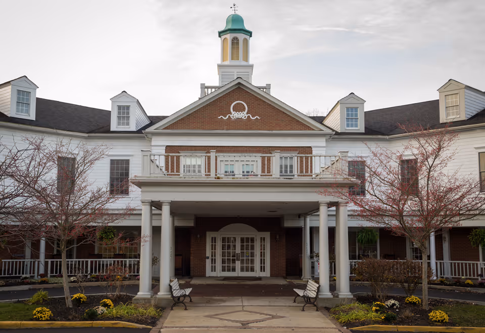Front exterior view of Poland Village Assisted Living building with a covered entrance supported by white columns, two benches, small trees with red leaves, and a cupola on the roof.