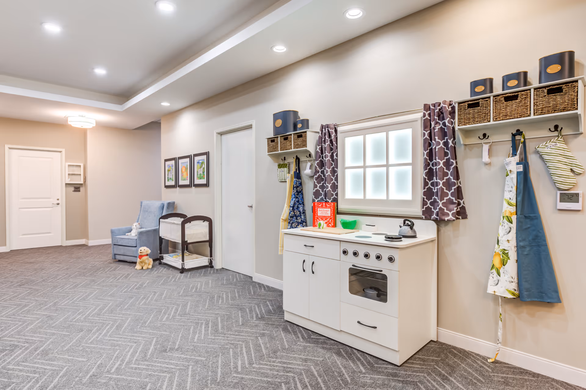 A bright and clean interior hallway area featuring a small play kitchen set with a window above it, aprons and oven mitts hanging on hooks, a blue armchair with a stuffed animal on it, a changing table, and framed pictures on the wall. The floor is carpeted with a herringbone pattern and the walls are painted light beige.