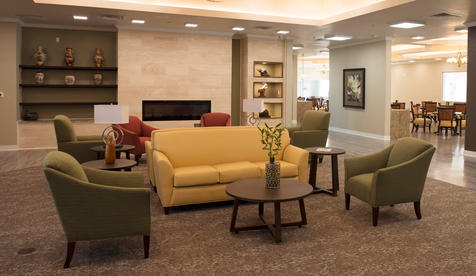 A senior living facility common area with a yellow sofa, four green armchairs, two red armchairs, and several round wooden tables. The room features a modern fireplace set into a tiled wall, decorative shelves with vases and sculptures, and a dining area visible in the background. The space is well-lit with ceiling lights and has a carpeted floor.
