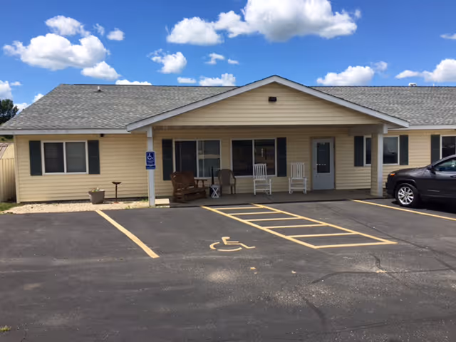 Single-story beige building front with a covered porch, rocking chairs, and marked handicap parking spaces.