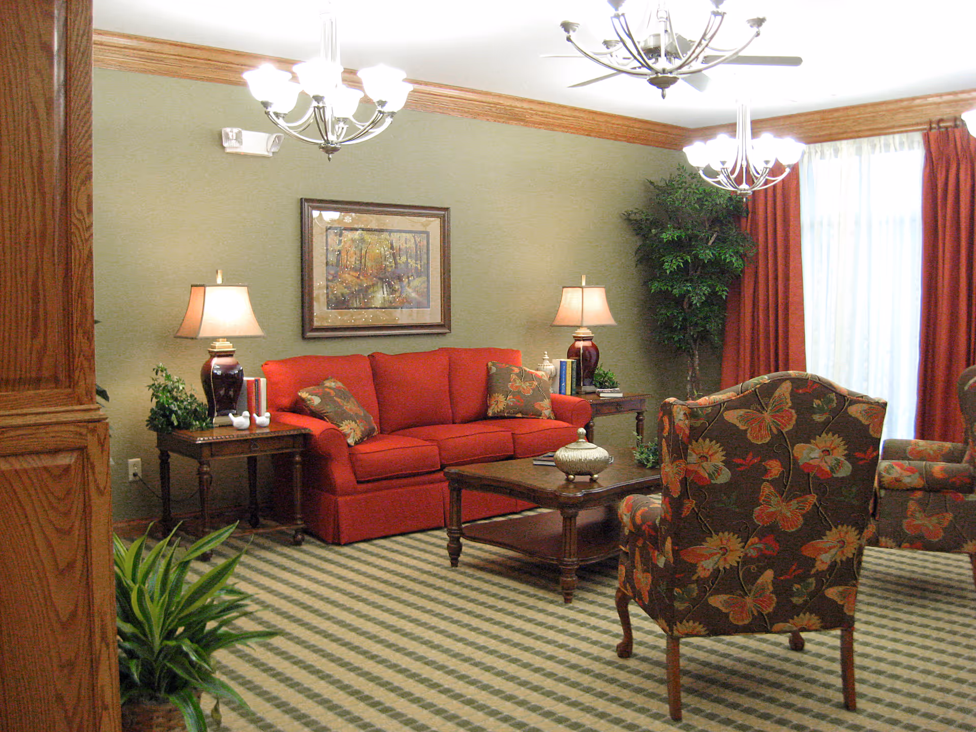 A cozy living room with a red sofa, two floral patterned armchairs, a wooden coffee table, side tables with lamps, a framed painting on the wall, potted plants, and large windows with red curtains.