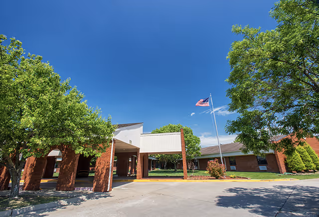 Exterior view of Carthage Health and Rehabilitation Center showing a brick building with a covered entrance, an American flag on a flagpole, and green trees under a clear blue sky.