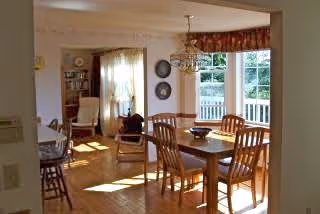Bright open dining area with a wooden table and chairs by a bay window overlooking a porch, adjacent to a living room with an armchair and bookcase.