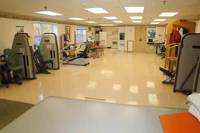 A spacious rehabilitation room with exercise equipment including stationary bikes, weight machines, and parallel bars. The room has a clean, polished floor, fluorescent ceiling lights, windows letting in natural light, and a small kitchenette area in the back with a refrigerator and oven. There are chairs and a table near the kitchenette, and various rehabilitation tools and posters on the walls.