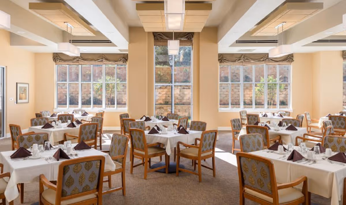 Bright dining room with tables set with white linens and folded napkins, surrounded by wooden chairs.