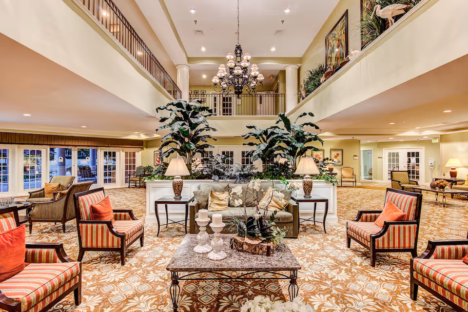 Spacious and elegant senior living facility common area with patterned carpet, multiple seating arrangements including striped armchairs with red cushions and a central sofa with beige pillows. The room features large indoor plants, two table lamps on side tables, a decorative coffee table with candles and floral arrangements, and a large chandelier hanging from the high ceiling. The area has a second-floor balcony overlooking the space and multiple doors leading outside.