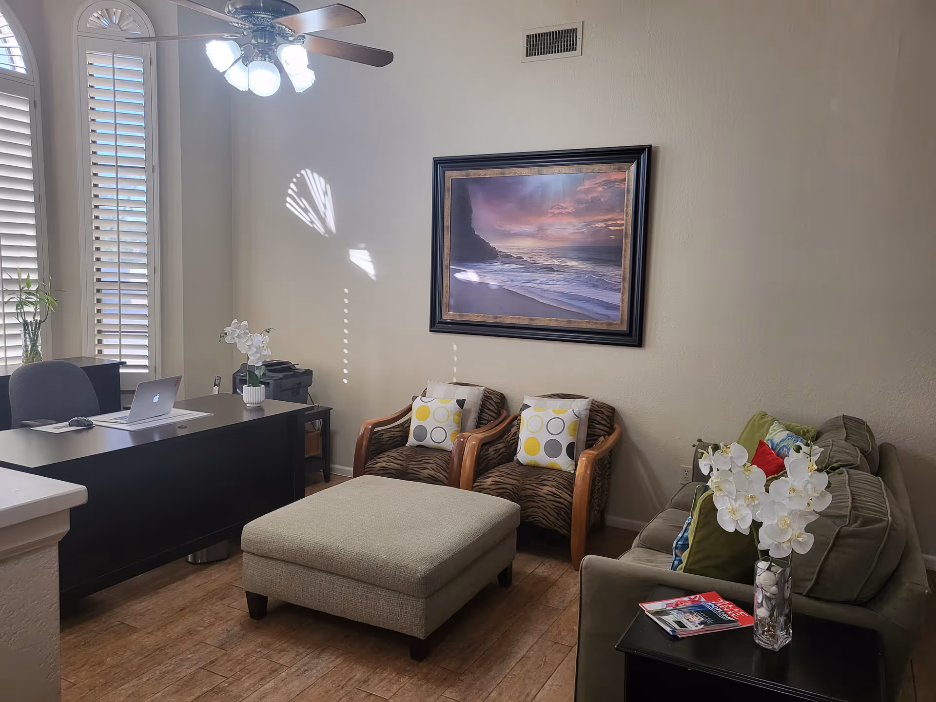 Sunlit reception-style living room with a desk, sofa, two chairs, ottoman, and framed beach artwork on the wall.