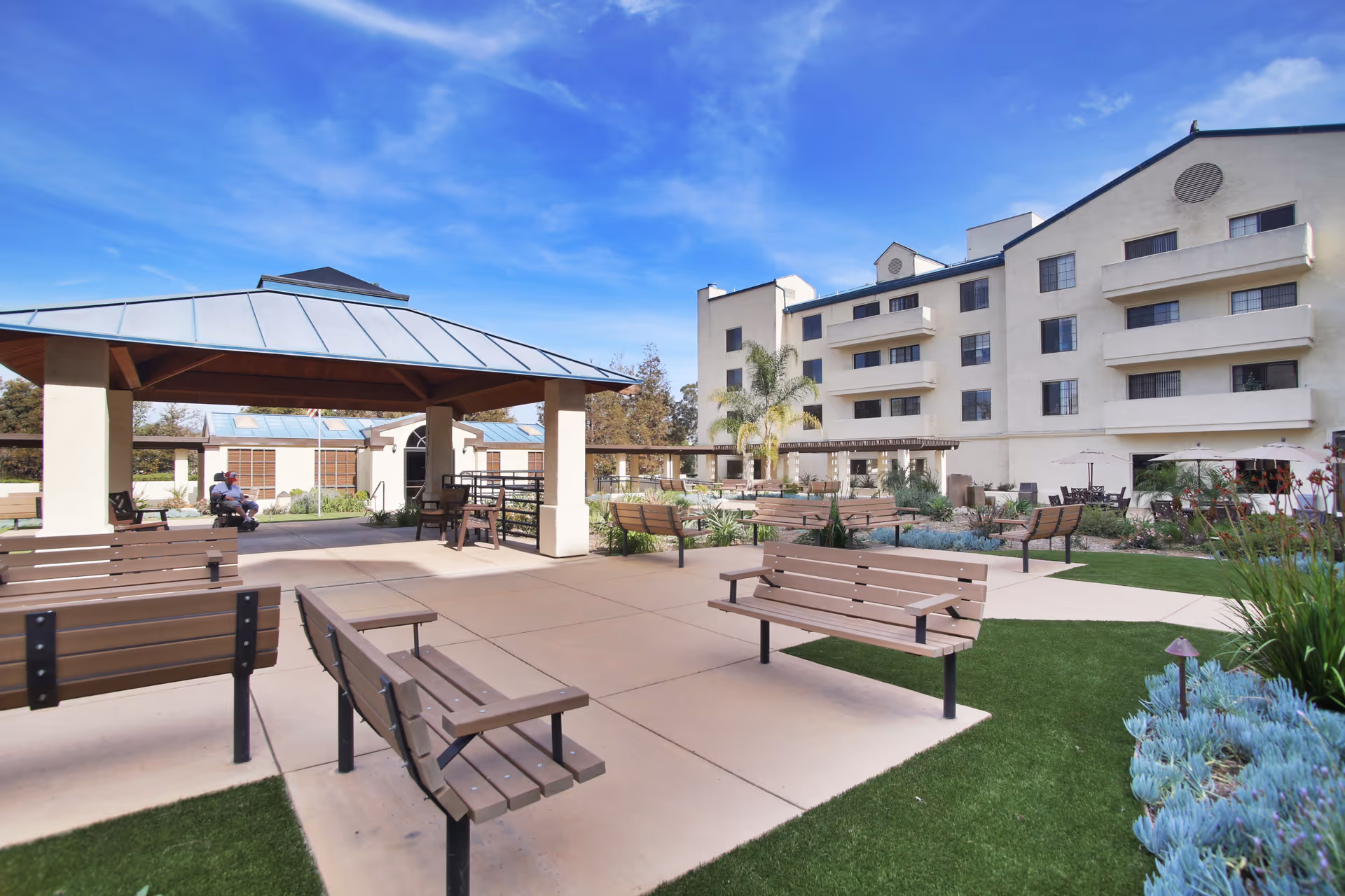 Outdoor seating area at Westmont of Escondido featuring multiple wooden benches on a paved patio with green grass and plants surrounding it. A covered pavilion with a metal roof is visible on the left side, and a multi-story residential building is in the background under a blue sky with some clouds.
