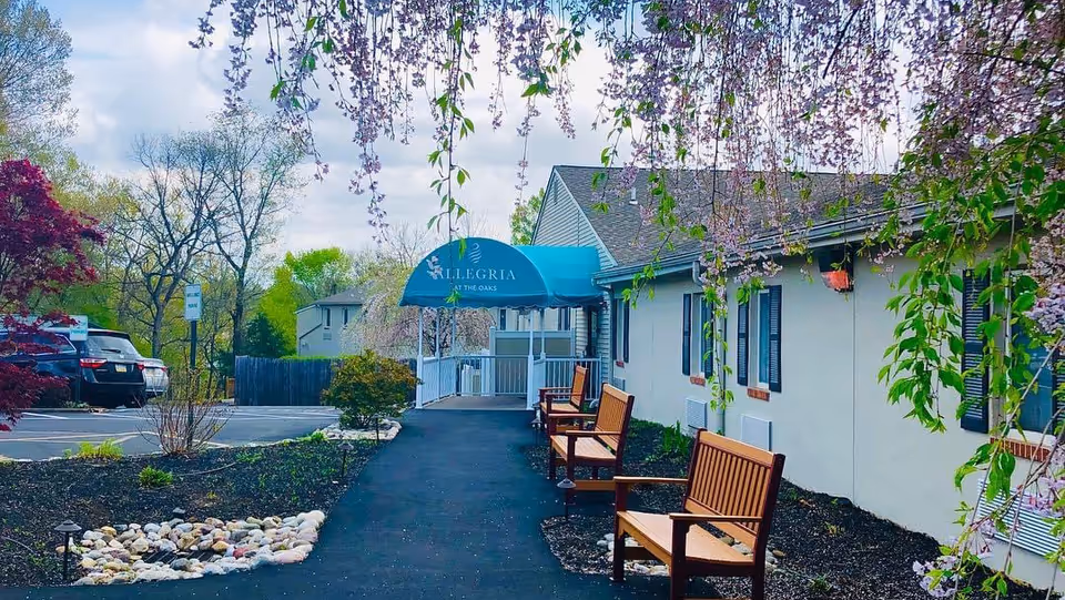 Exterior view of Allegria at The Oaks facility entrance with a blue awning displaying the facility name. There is a paved walkway leading to the entrance, flanked by wooden benches and landscaped garden beds with rocks and plants. Trees with hanging flowers frame the top of the image, and parked cars are visible in the background.