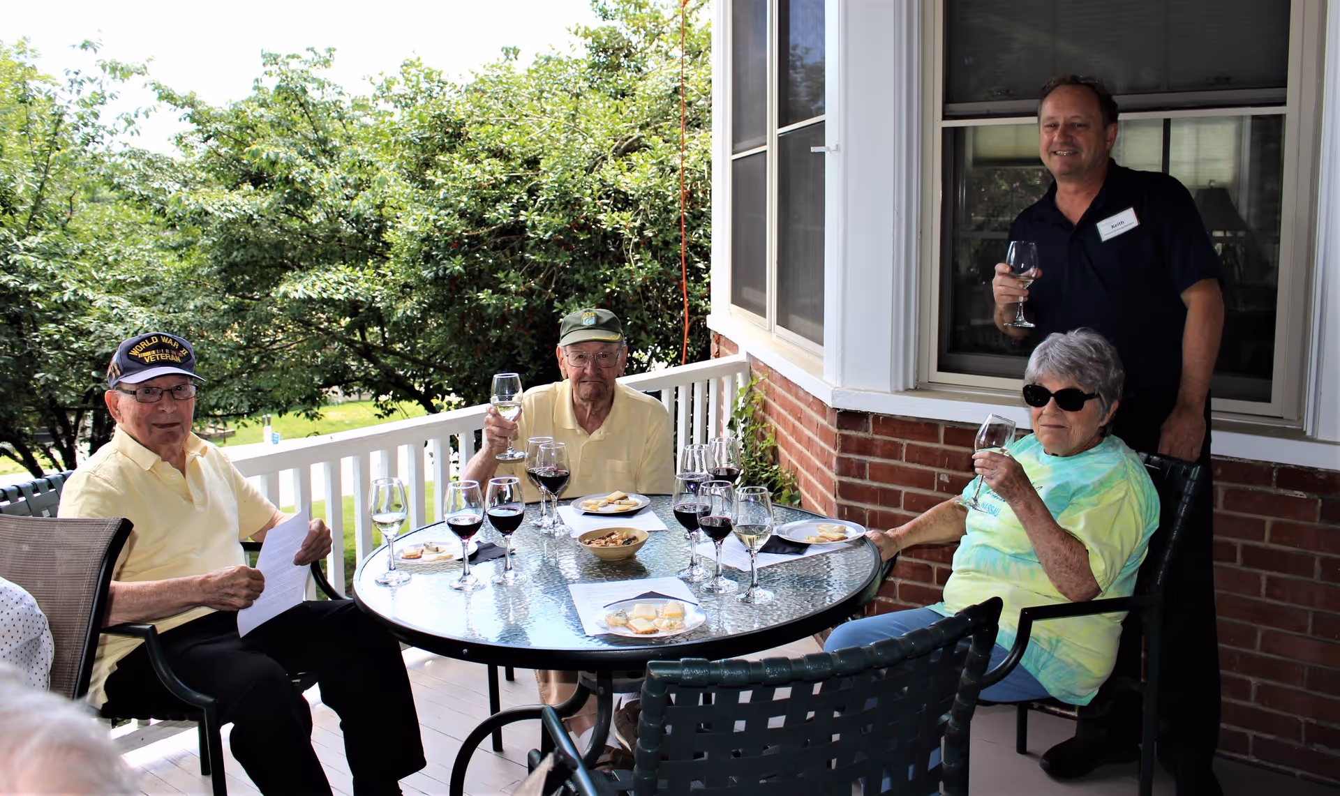 Four elderly individuals sitting around a glass outdoor table on a porch, enjoying glasses of wine and snacks. The setting is bright with greenery in the background, and one man is standing behind the seated group, smiling and holding a wine glass.