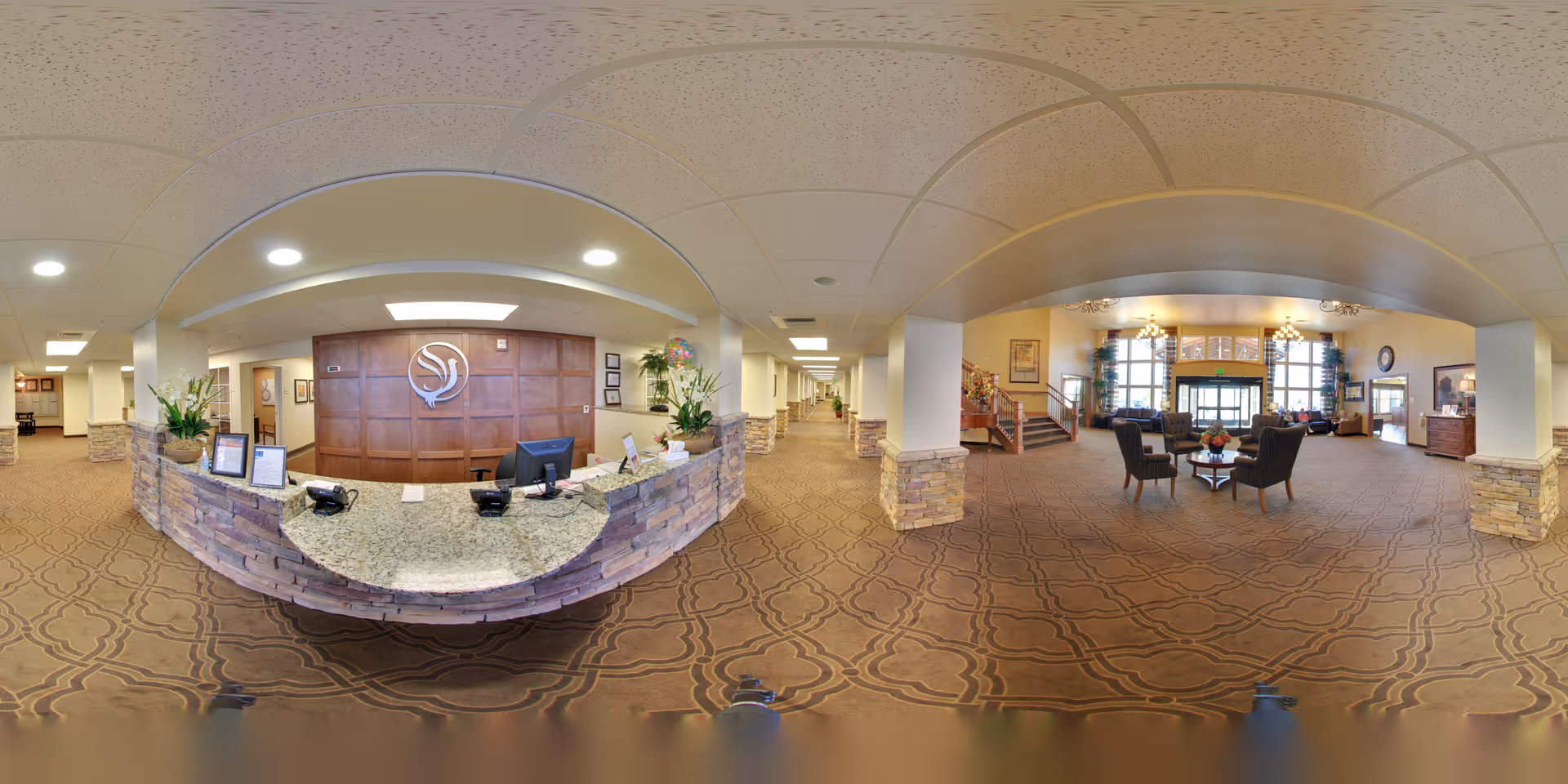 Spacious senior living facility lobby with a curved stone reception desk on the left and a seating area with chairs and tables on the right under high ceilings.