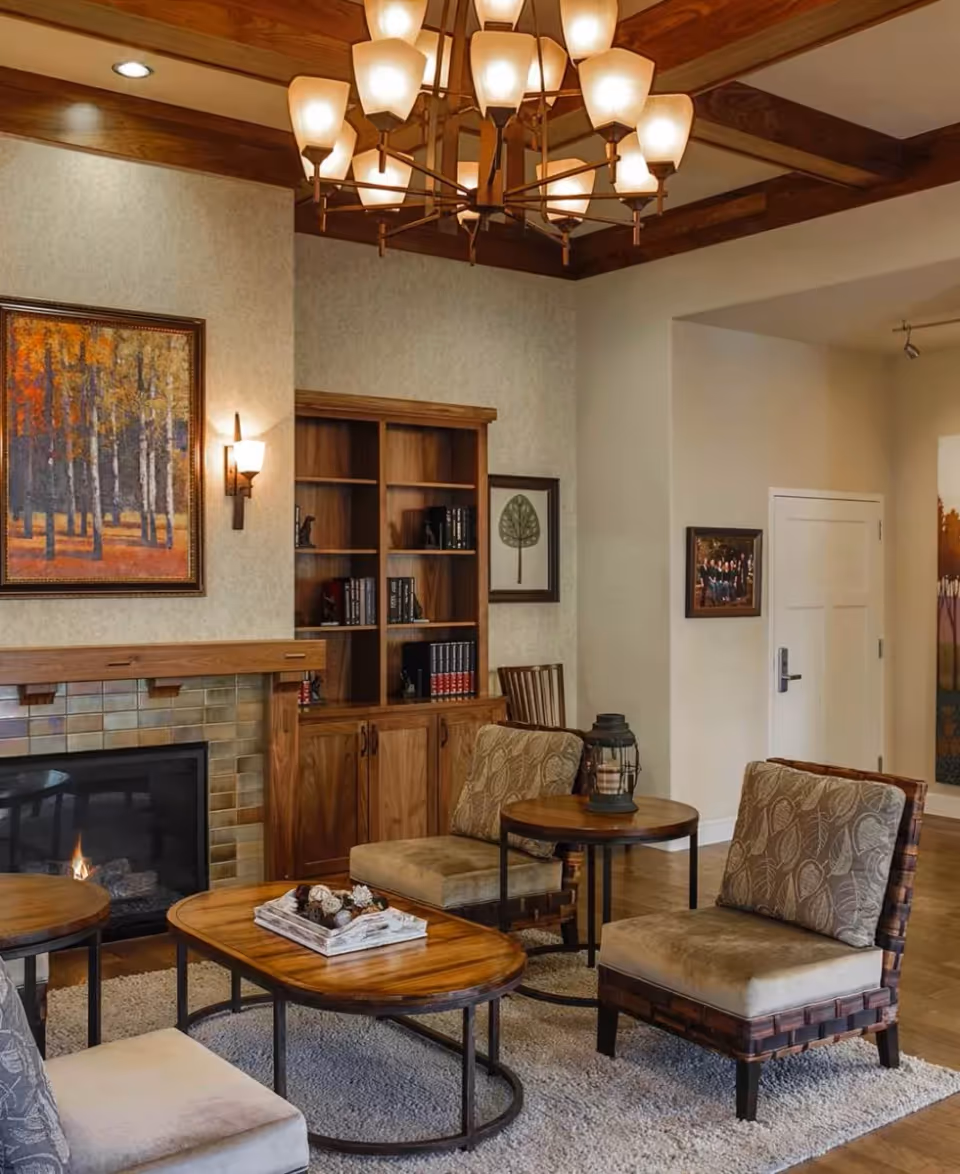 A cozy living room area with a wooden ceiling featuring exposed beams and a large chandelier. The room includes a fireplace with a tiled surround, a wooden bookshelf with books and decor, two cushioned chairs with patterned pillows, and wooden coffee and side tables. The walls are decorated with framed artwork, and the floor is covered with a light-colored rug.