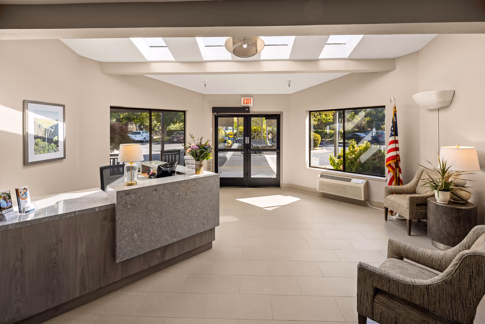 Reception area of Provincial Chico Senior Living with a modern front desk, two large windows, an exit door, two armchairs, a side table with a lamp and plant, an American flag, and a framed picture on the wall. The space is well-lit with natural light from skylights.