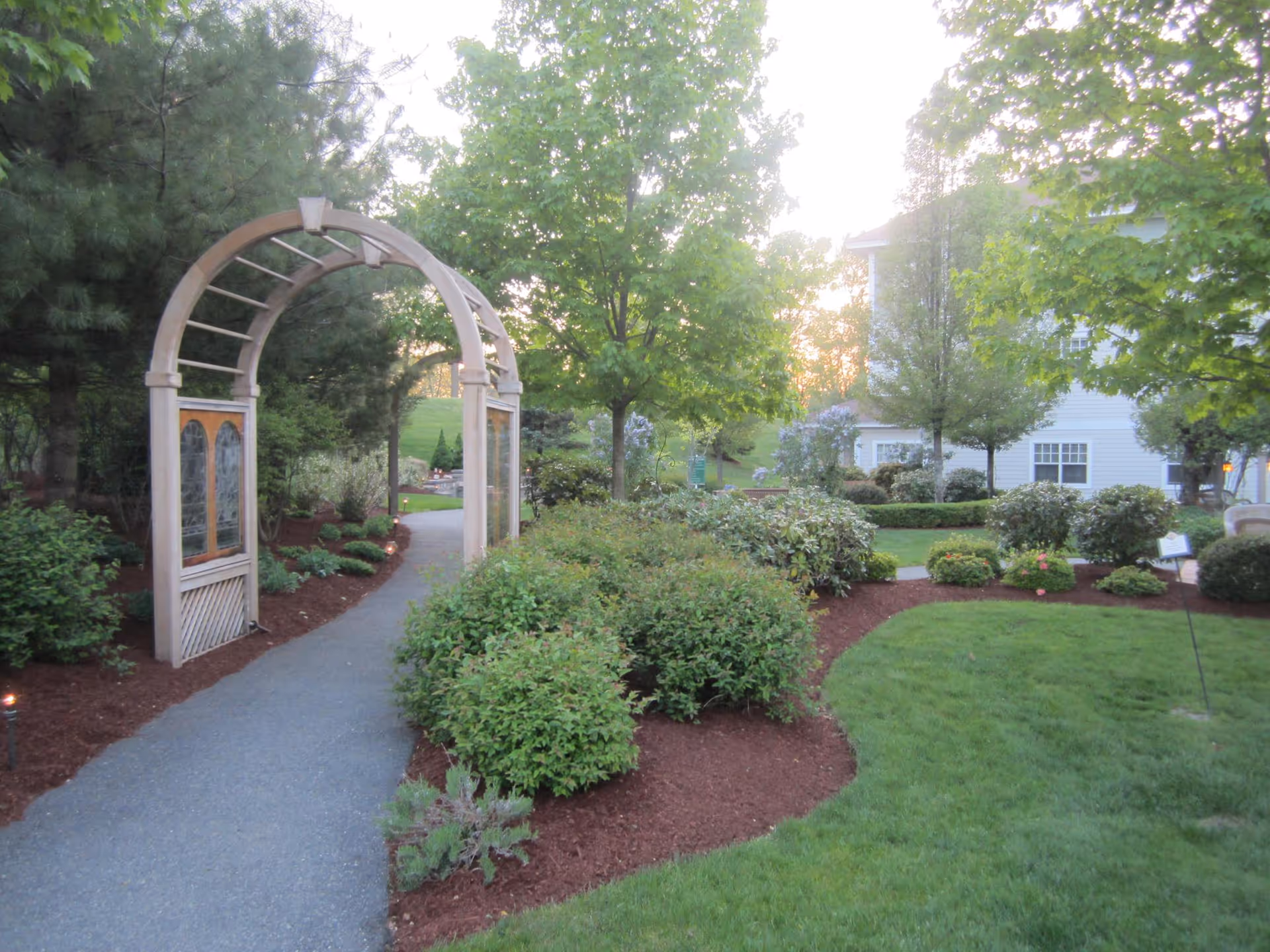 A landscaped garden path at Tamarisk Senior Living featuring a wooden archway with decorative panels, surrounded by green bushes, trees, and neatly mulched flower beds. A white building is visible in the background with sunlight filtering through the trees.