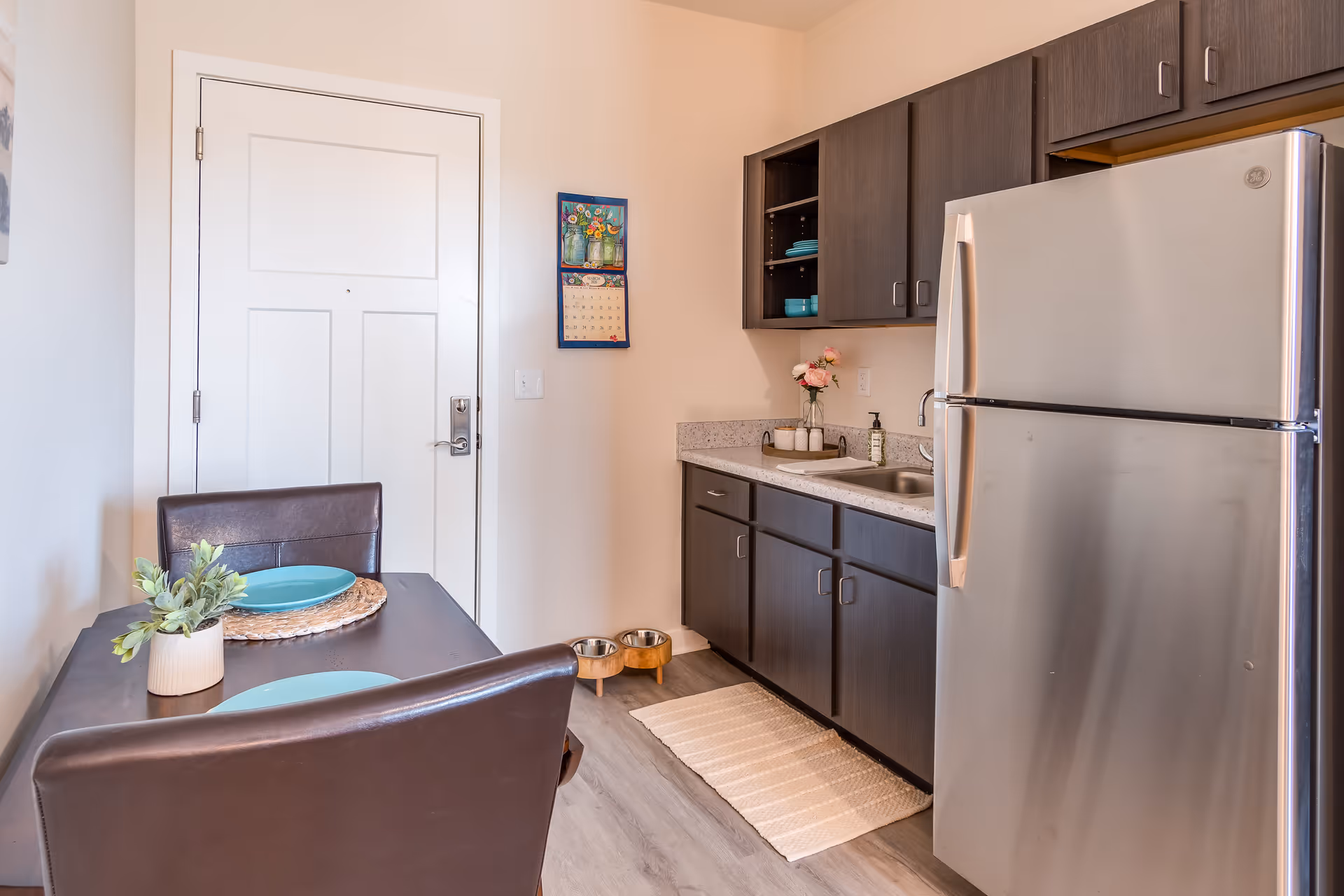 A small kitchen area with dark wood cabinets, a stainless steel refrigerator, a countertop with a sink, and a small dining table set for two with blue plates and a small plant. A white door is visible in the background along with a wall calendar and two pet bowls on the floor.