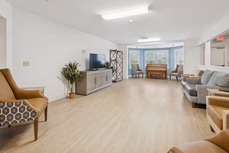A spacious, well-lit living room with light wood flooring and white walls. The room features a TV on a wooden cabinet, several armchairs and sofas arranged along the walls, a wooden piano in a bay window area with three chairs, and some potted plants for decoration.