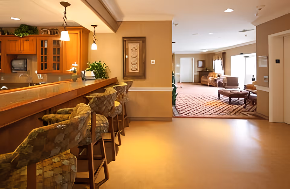 Interior view of a senior living facility showing a bar area with high chairs on the left and a lounge area with sofas and a coffee table in the background. The space is warmly lit with pendant lights above the bar and natural light coming from windows in the lounge area.