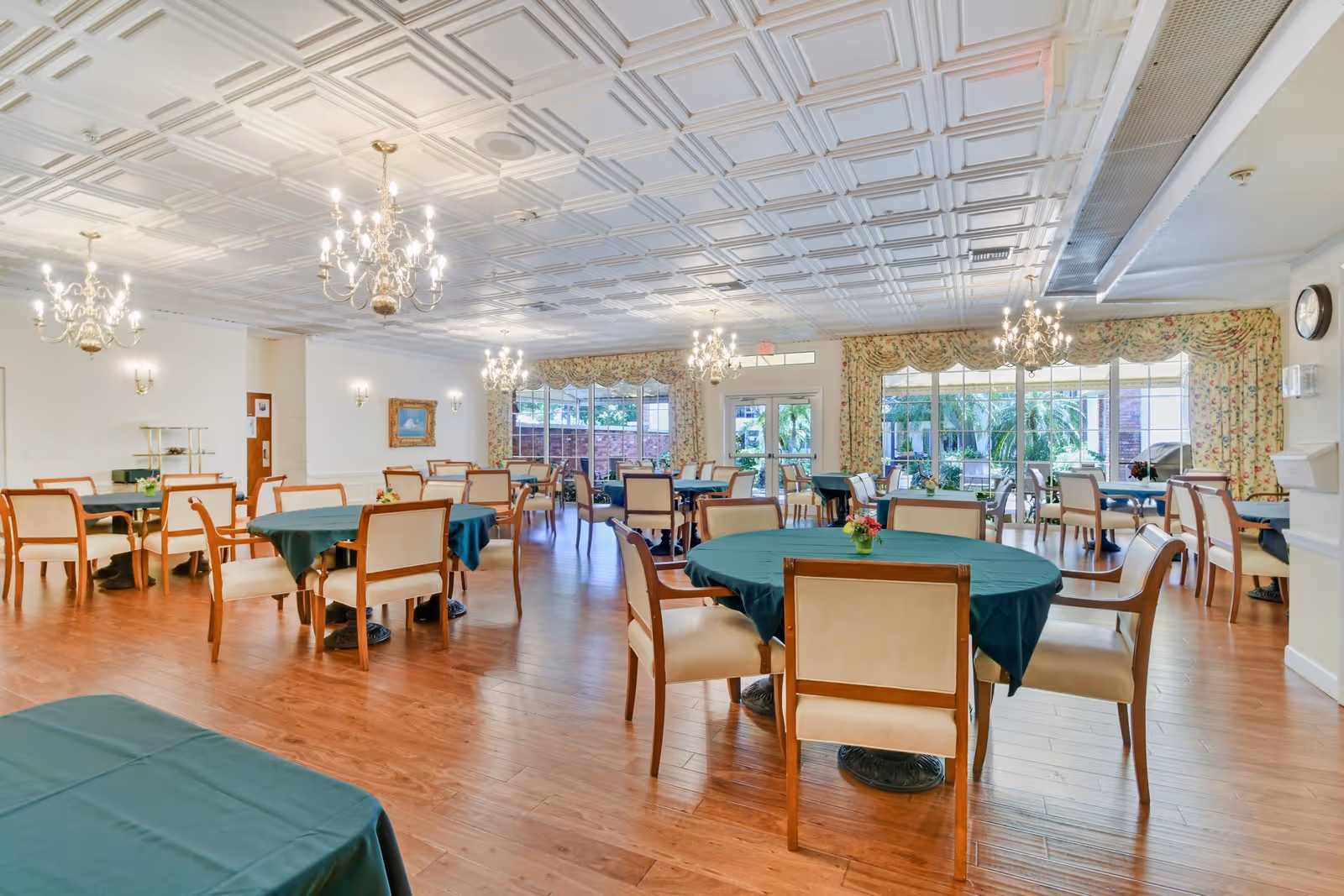 Spacious assisted living dining room with round tables draped in green tablecloths, wooden chairs, chandeliers, and large windows with floral curtains.