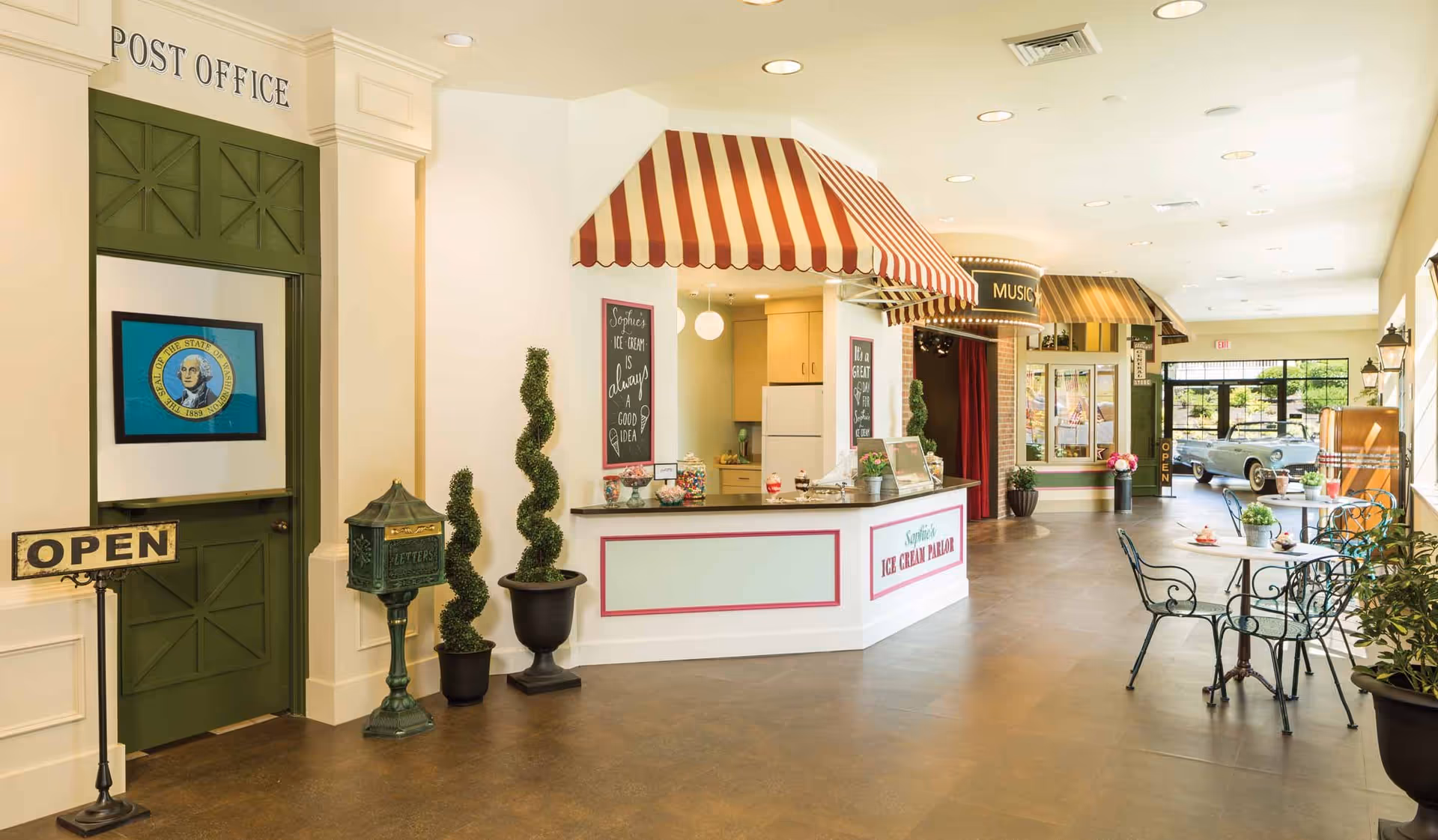 Interior view of a senior living facility area designed to resemble a small town street with a post office, an ice cream parlor with a red and white striped awning, and a music store. There are small tables and chairs with plants and desserts on them, decorative topiary plants, and a vintage car visible near large windows letting in natural light.