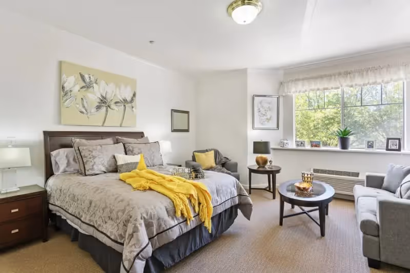 A bright and cozy bedroom featuring a large bed with gray floral bedding and a yellow throw blanket. The room includes two bedside tables with lamps, a comfortable armchair with a yellow pillow, a round coffee table, and a gray sofa near a large window with a view of green trees. The walls are decorated with floral artwork and a mirror.