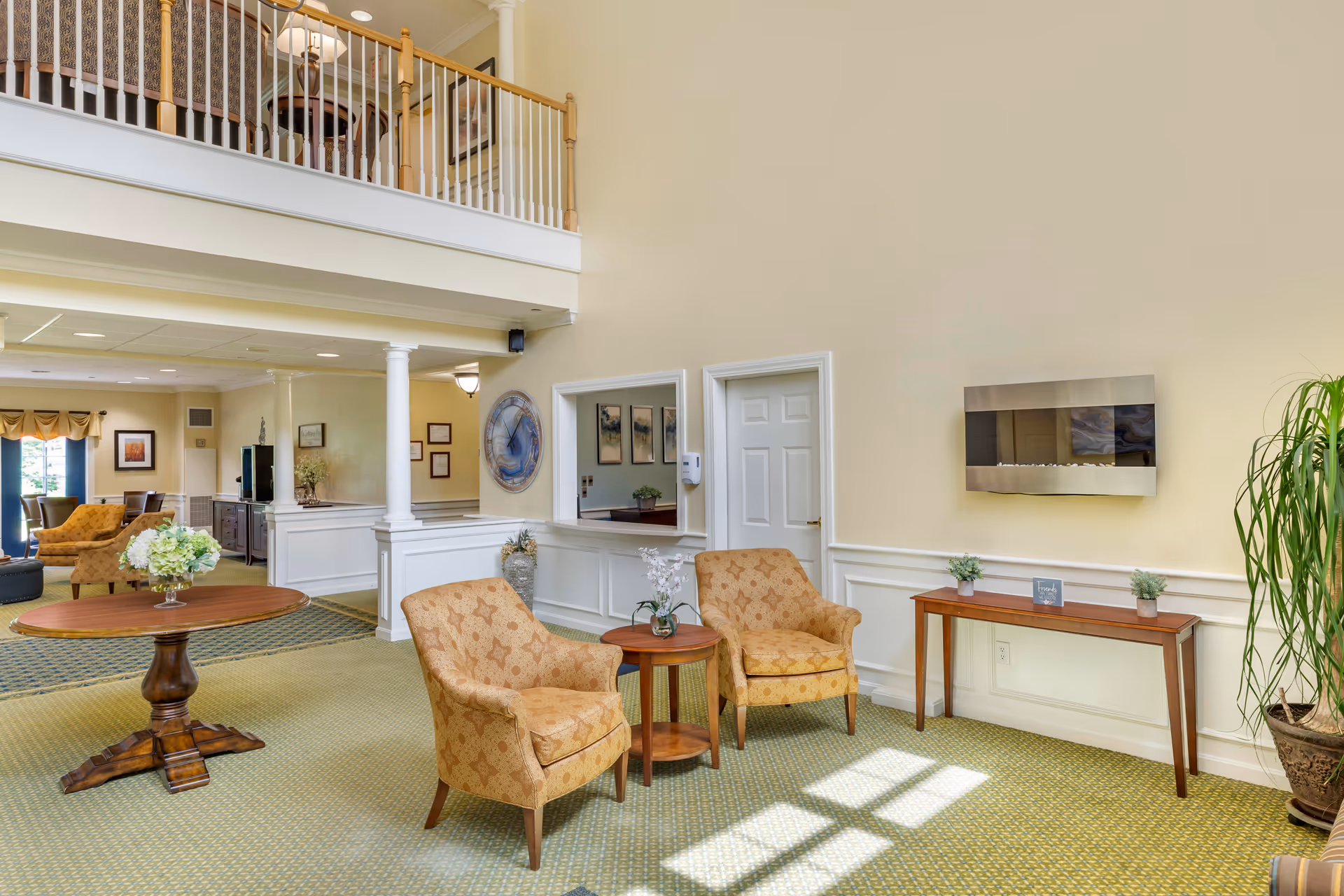 A bright and spacious senior living facility common area with two patterned armchairs and a small round wooden table between them. There is a wooden console table against the wall with small plants and a decorative item. A modern wall-mounted electric fireplace is above the console table. The room has light yellow walls, white trim, and green patterned carpet. A large round wooden table with a flower arrangement is visible to the left, and there is a balcony railing above the seating area.