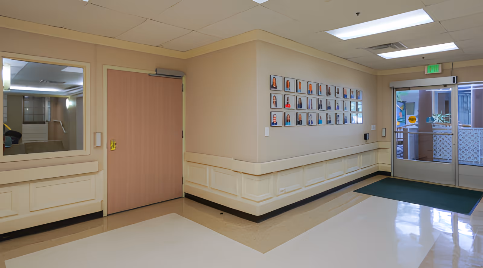 Interior hallway of Hale Nani Rehab & Nursing Center with beige walls, a wooden door, a large window on the left, a bulletin board with multiple framed photos on the right wall, and glass double doors leading outside at the end of the hallway.