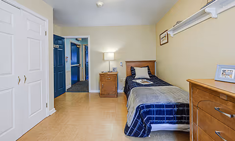 A simple bedroom with a single bed covered in a blue and gray blanket, a wooden nightstand with a lamp, a wooden dresser with a framed photo, and a closet with white double doors. The walls are light yellow, and there is a blue door leading to another room.