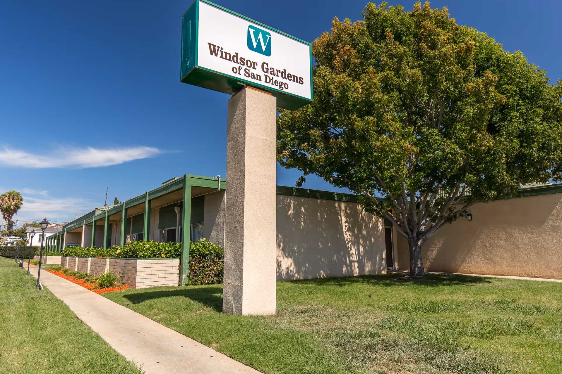 Exterior front of Windsor Gardens Convalescent Center of San Diego with a large sign, building facade, sidewalk, lawn, and a tree.