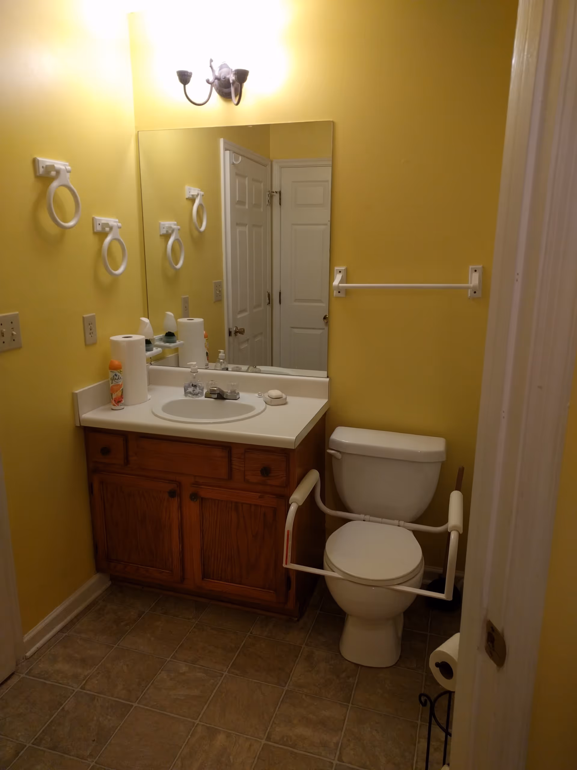 A small bathroom with yellow walls featuring a white toilet equipped with safety rails, a wooden vanity with a white countertop and sink, a large mirror above the sink, a wall-mounted light fixture, and several towel holders. The floor is tiled in a brown pattern.