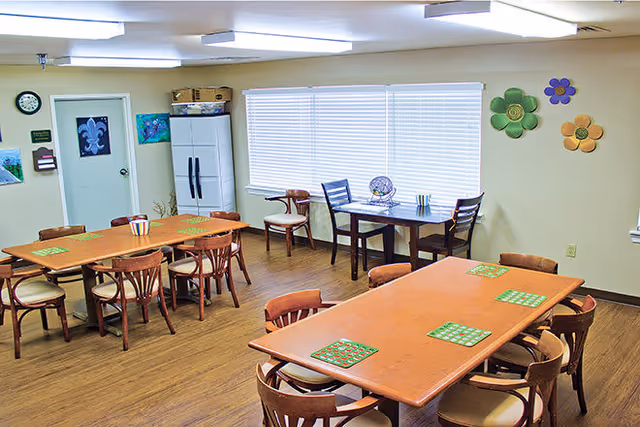 A well-lit room with wooden tables and chairs arranged for group seating. The tables have green placemats, and there is a bingo cage and cards on one table near the window with closed blinds. The walls are decorated with colorful flower-shaped wall hangings, and there is a clock and some artwork on the far wall. A white refrigerator is also visible in the corner.