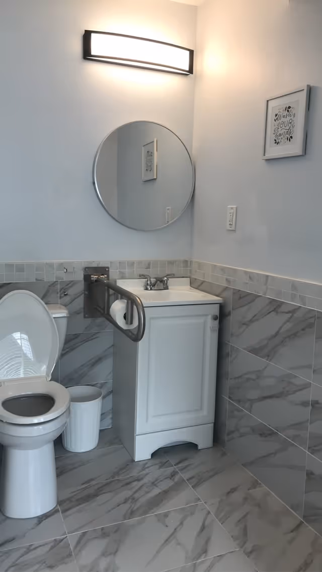 A clean bathroom with a white toilet, a white sink cabinet with a silver faucet, a round mirror above the sink, a wall-mounted light fixture, a handrail with a toilet paper holder attached, and a small white trash bin on the floor. The walls are tiled halfway up with gray marble tiles and the upper half is painted white. A framed sign on the wall reads 'Wash Your Hands'.