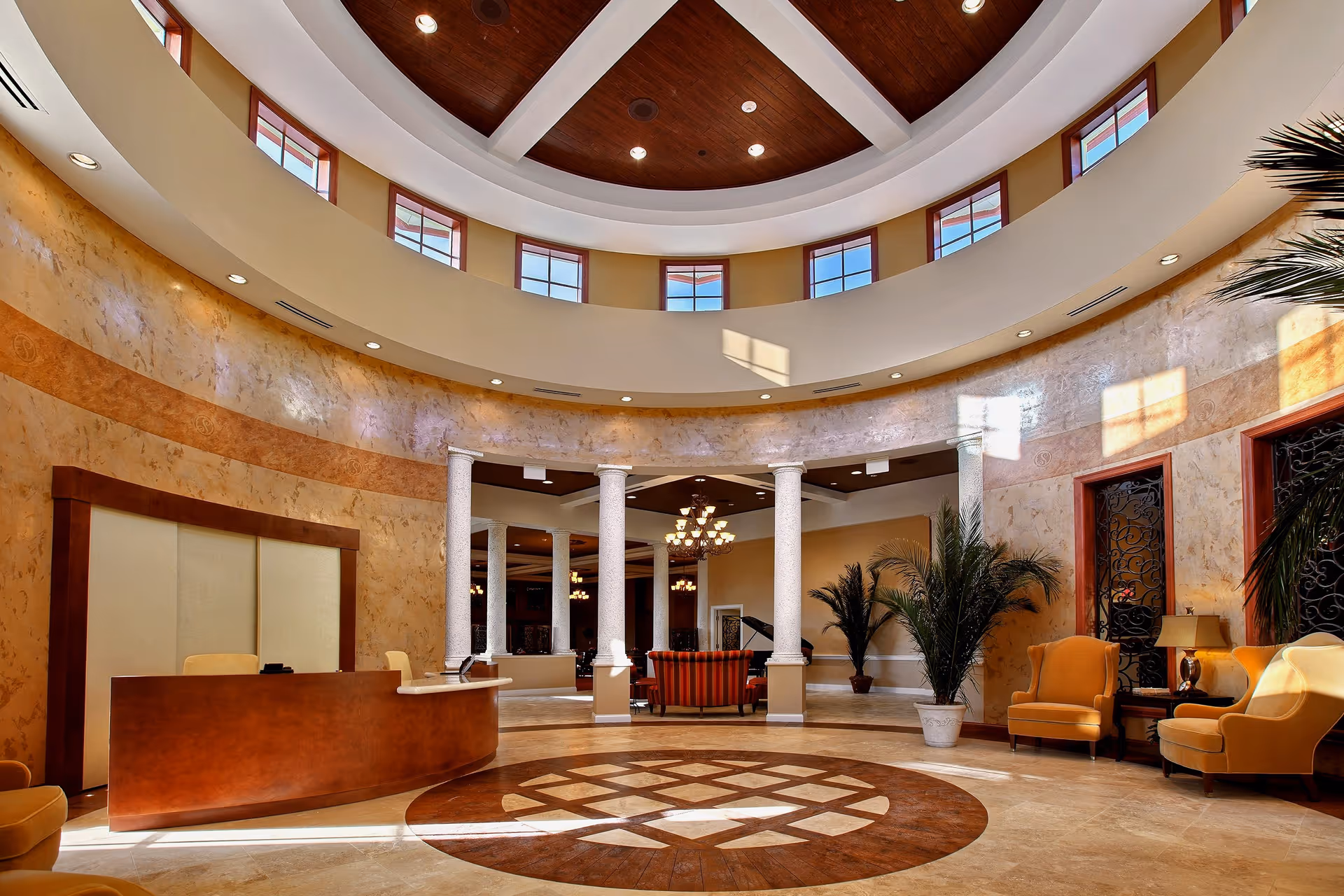 A spacious, elegant lobby area with a curved reception desk, marble flooring with a decorative circular pattern, tall white columns, and a high ceiling with multiple small windows near the top. The room is warmly lit with chandeliers and natural light, and there are comfortable armchairs and potted plants arranged around the space.