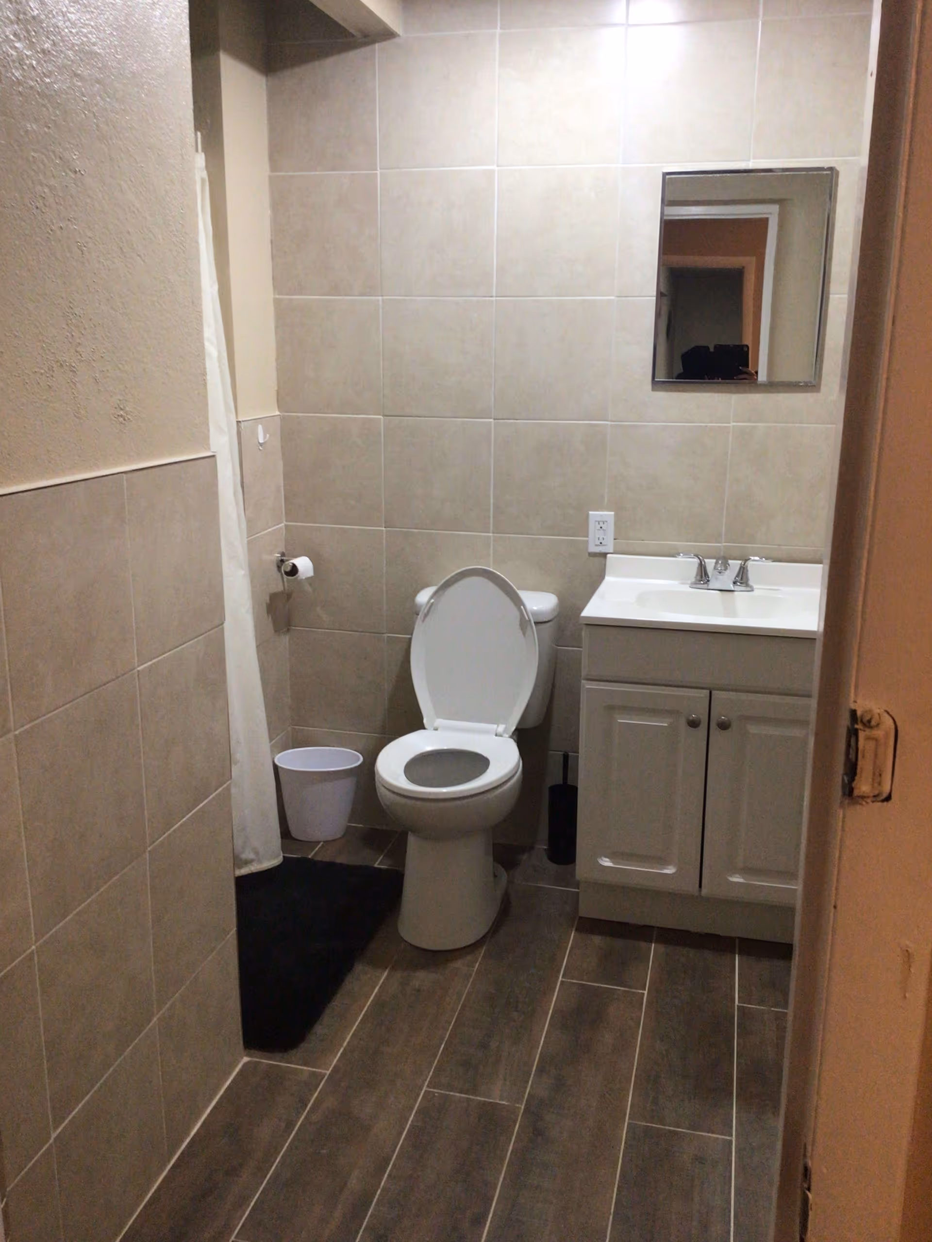 A small bathroom with beige tiled walls and wood-look tile flooring. The bathroom features a white toilet with the lid open, a white vanity with a sink and faucet, a rectangular mirror above the sink, a black bath mat, a white trash bin, and a toilet paper holder mounted on the wall.