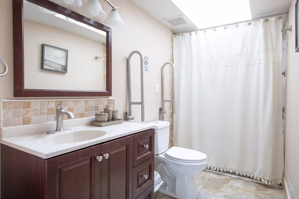 A clean and well-lit bathroom featuring a dark wood vanity with a white countertop and sink, a large mirror with a dark wood frame above the sink, a toilet with safety grab bars on either side, and a shower with a white curtain. The walls are light-colored with a tiled backsplash behind the sink and tiled flooring.