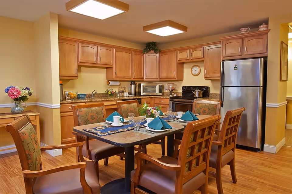 A warm and inviting kitchen and dining area with wooden cabinets, a stainless steel refrigerator, a stove, microwave, and coffee maker. The dining table is set with plates, cups, glasses, and teal napkins, surrounded by six wooden chairs with floral upholstery. A vase with flowers is placed on a side table to the left.