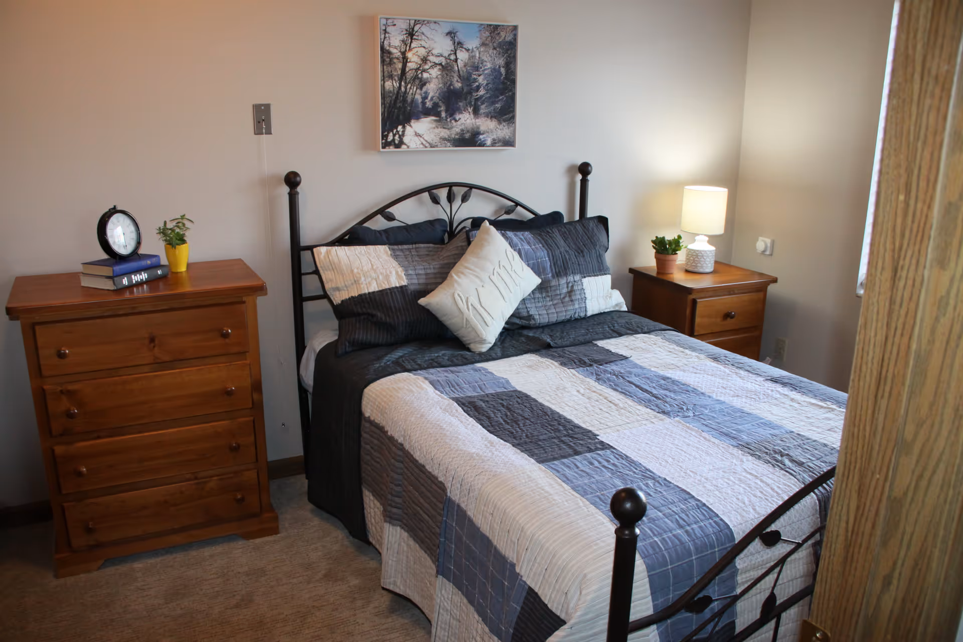A cozy bedroom with a metal framed bed covered in a patchwork quilt in shades of blue, gray, and white. There are two wooden nightstands on either side of the bed, one with a clock and small plant, and the other with a lamp and a small potted plant. A framed picture of a snowy forest scene hangs on the wall above the bed.
