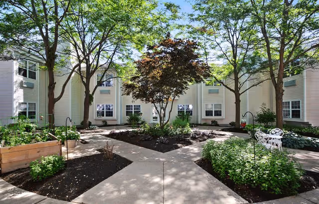 A peaceful outdoor courtyard at Shawnee Estates Senior Living featuring a paved walkway intersecting through garden beds with various plants and trees, surrounded by a two-story building with multiple windows.
