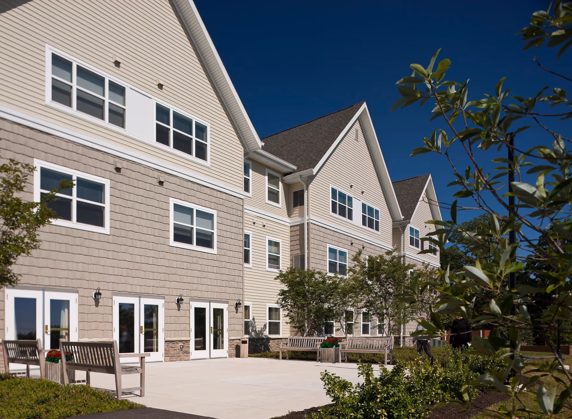 Exterior view of a senior living facility building with beige siding and multiple windows under a clear blue sky. There are several wooden benches and small trees planted along a paved patio area in front of the building.