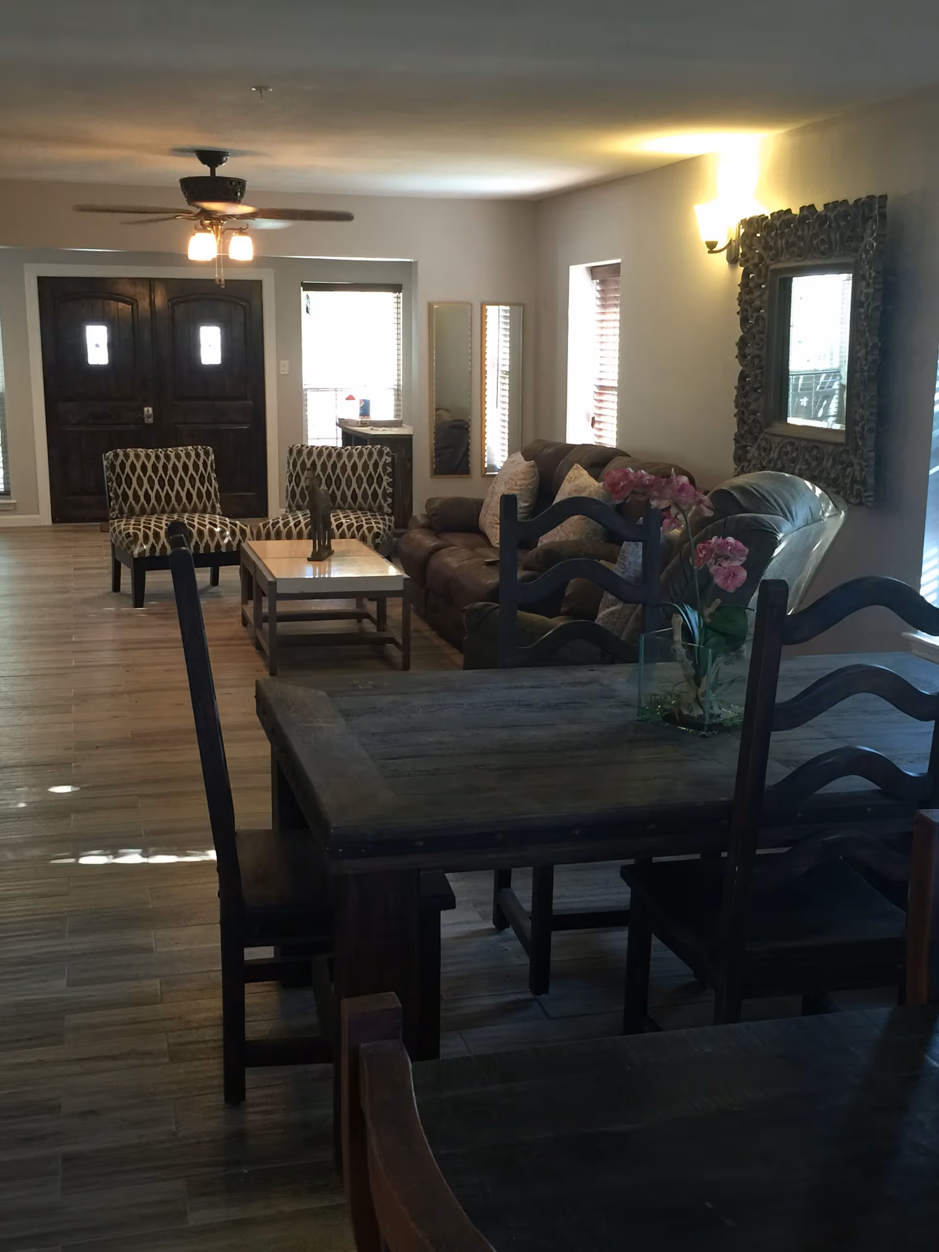 Interior view of a living room and dining area with wooden floors. The living room has a brown leather sofa, two patterned armchairs, a coffee table, and a large decorative mirror on the wall. The dining area has a dark wooden table with chairs and a vase with pink flowers on the table. There is a ceiling fan with lights and large double doors in the background.