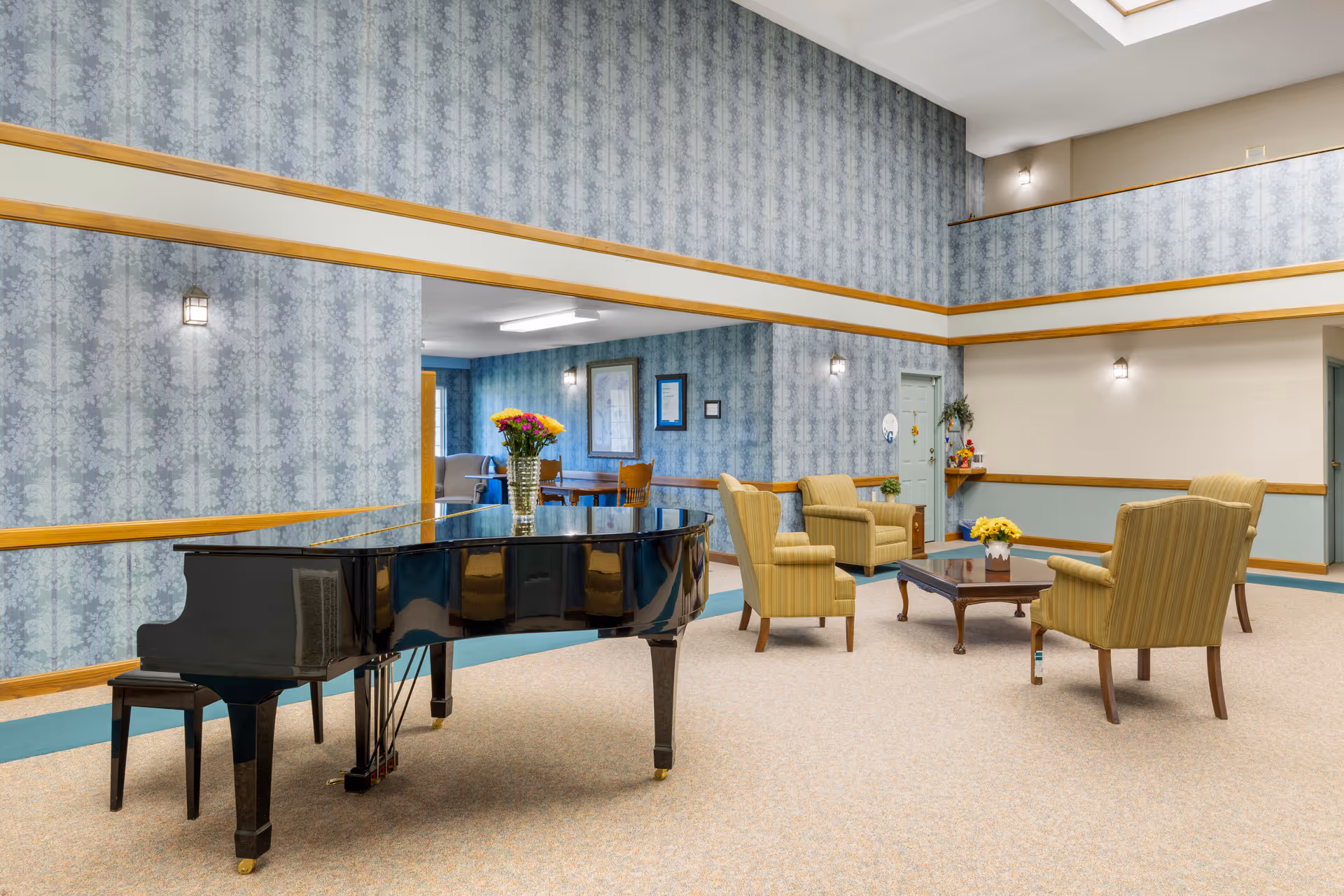 A spacious common area in a senior living facility featuring a black grand piano with a vase of flowers on top, four striped armchairs arranged around a wooden coffee table with a small flower arrangement, and blue patterned wallpaper with wooden trim. The room has a high ceiling with a balcony railing visible above and soft lighting fixtures on the walls.