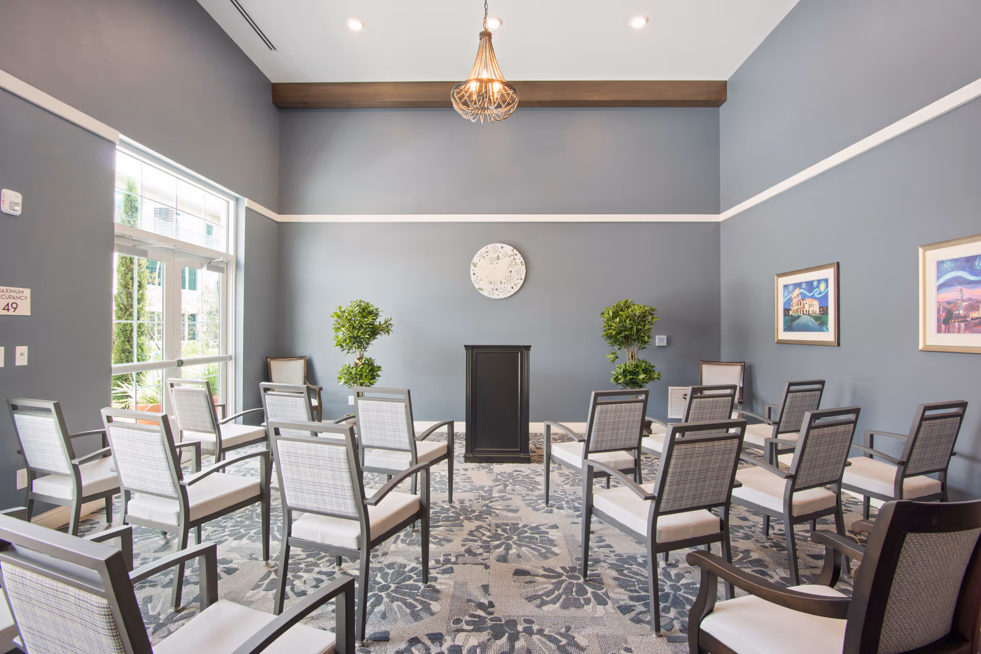 A small conference or meeting room with multiple chairs arranged in rows facing a black podium. The room has gray walls with white trim, a patterned carpet, two potted plants on either side of the podium, a round decorative wall piece above the podium, and two framed pictures on the right wall. There is a glass door on the left side letting in natural light.