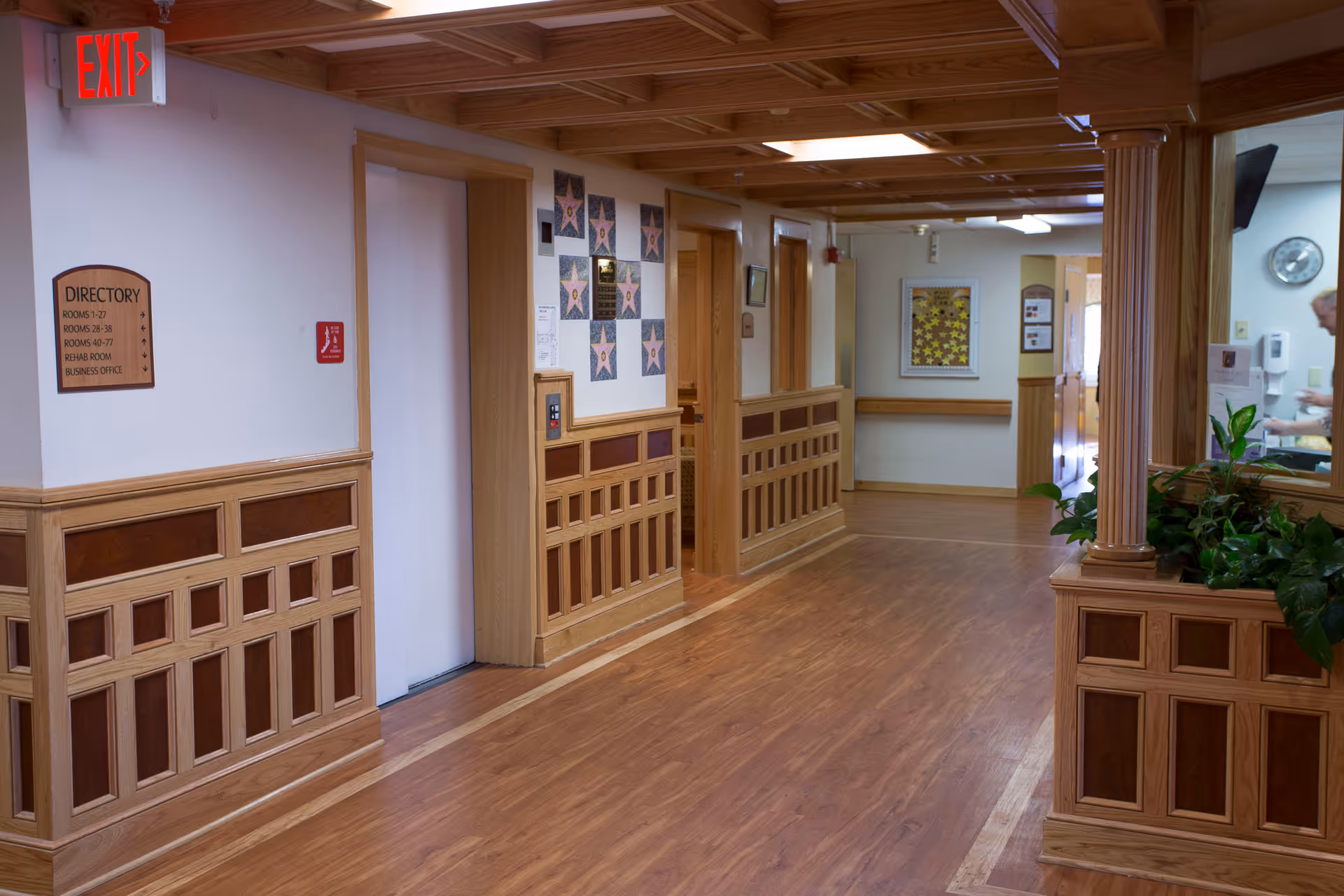 A clean, well-lit hallway in a senior living facility with wooden paneling and flooring. There is a directory sign on the left wall listing room numbers and offices, an exit sign above a door, and decorative star plaques on the wall. A person is partially visible in the background near a counter with plants.