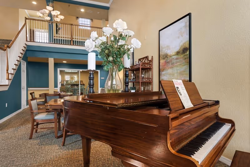 Wooden grand piano with a vase of white flowers in a spacious two-story common room with dining tables and an upper balcony.