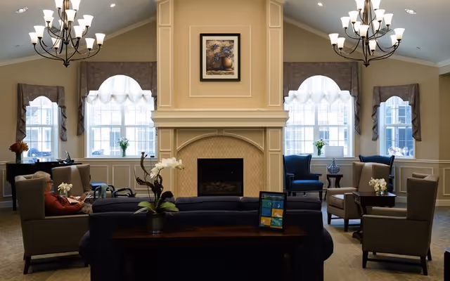 A cozy and elegant living room area with a central fireplace, flanked by two large windows with decorative curtains. The room features comfortable armchairs and sofas arranged around the fireplace, with small tables holding flower arrangements. Two chandeliers hang from the ceiling, providing warm lighting.