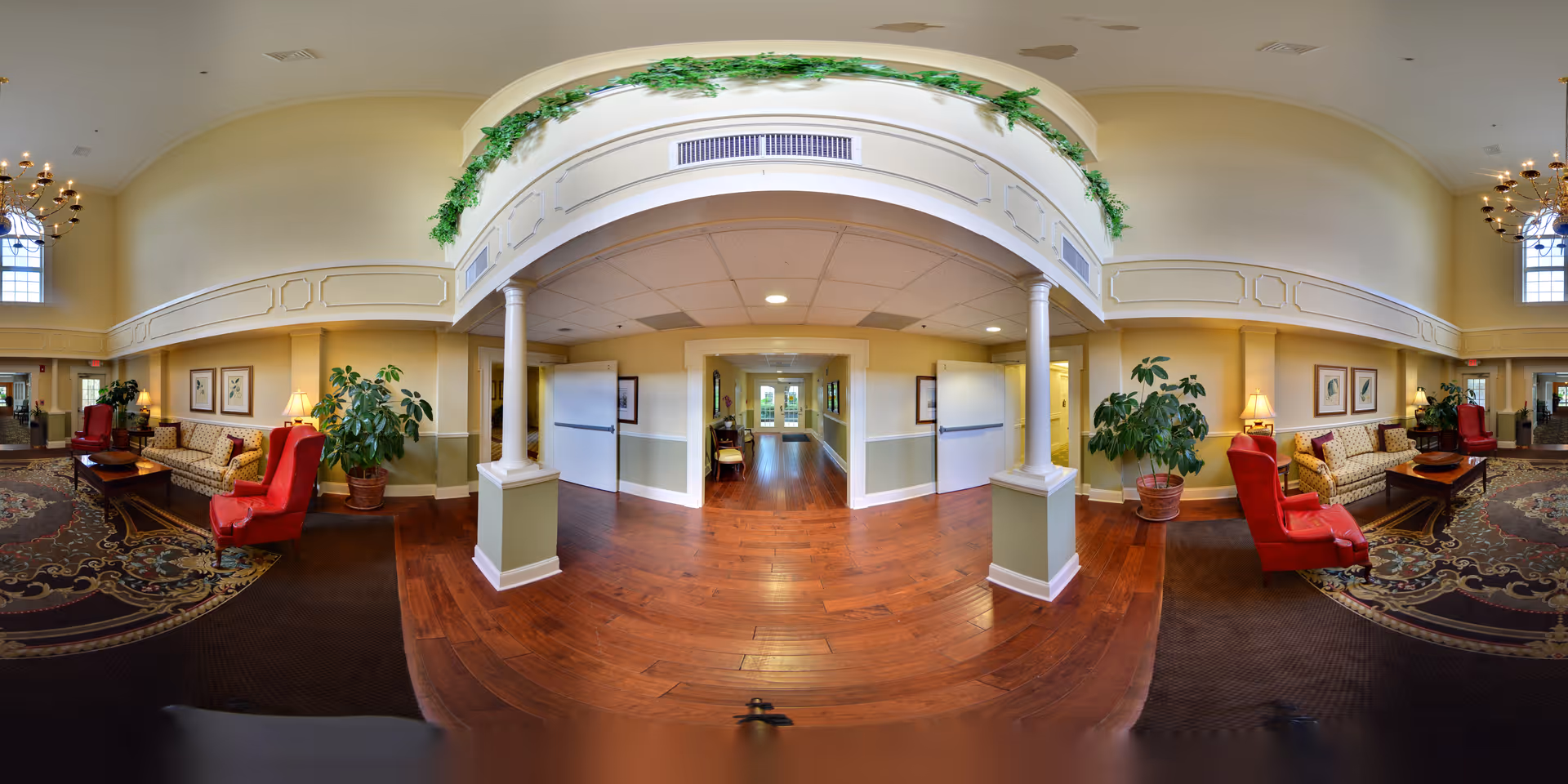 A spacious and well-lit common area in a senior living facility featuring comfortable seating with red armchairs and patterned sofas, decorative plants, framed artwork on the walls, and a large ornate rug on the floor. The area has high ceilings with chandeliers and an archway adorned with greenery leading to a hallway with wooden flooring.