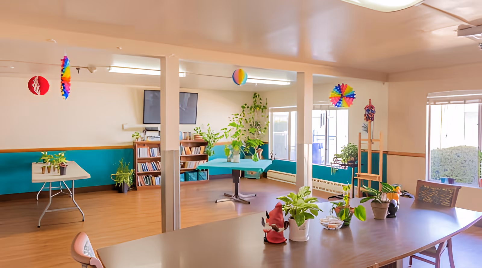 A bright and spacious common area in a nursing center with wooden floors and teal lower wall panels. The room features a large table with potted plants and decorative items, a smaller table with more plants, a bookshelf filled with books, and colorful hanging decorations from the ceiling. Large windows allow natural light to fill the space.
