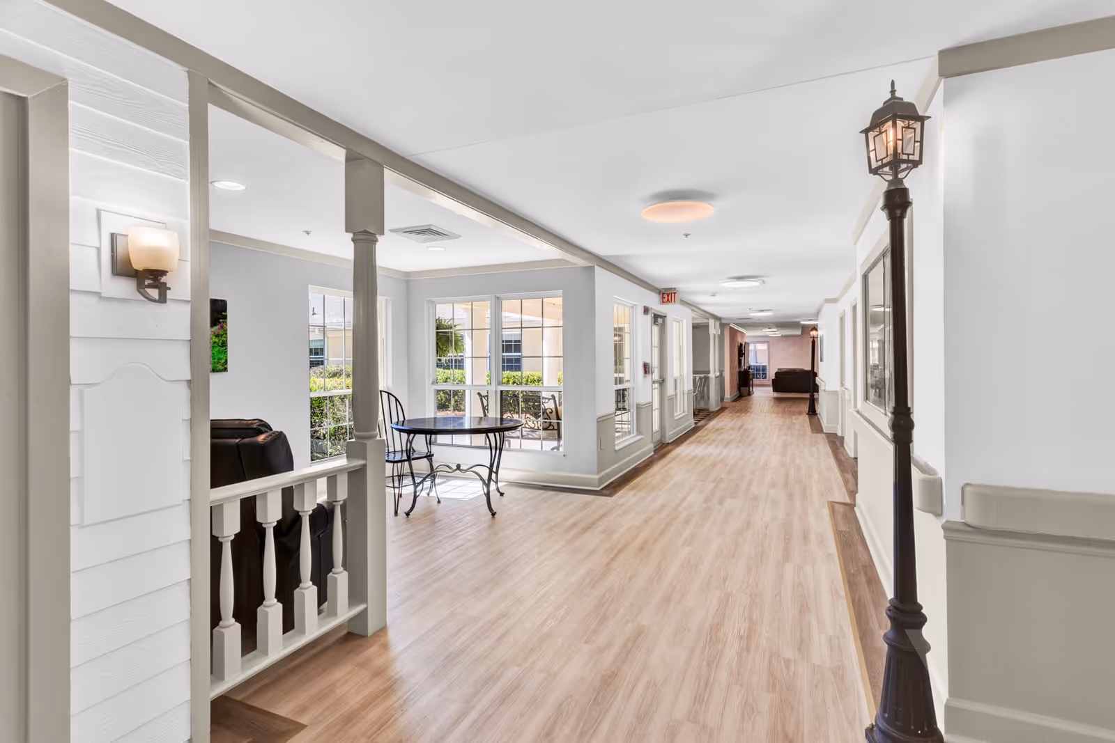 A bright and spacious hallway in an assisted living facility with light wood flooring, white walls, and beige trim. The hallway features large windows letting in natural light, a small round table with two chairs near the windows, and a decorative street lamp-style light fixture along the wall. There are seating areas with dark leather chairs visible in the background.
