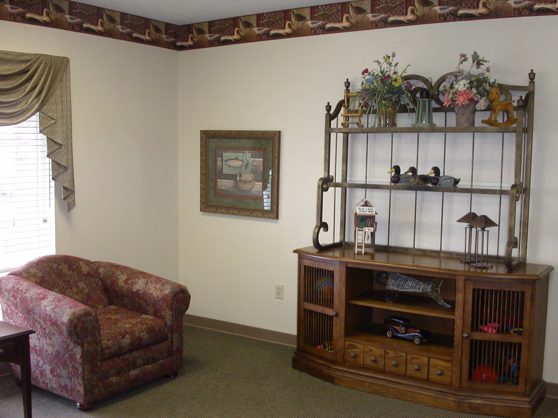 A cozy corner of a room featuring a floral upholstered armchair next to a window with draped curtains. On the adjacent wall, there is a framed picture. A wooden cabinet with a metal rack on top holds decorative items including flowers, small bird figurines, and other ornaments.