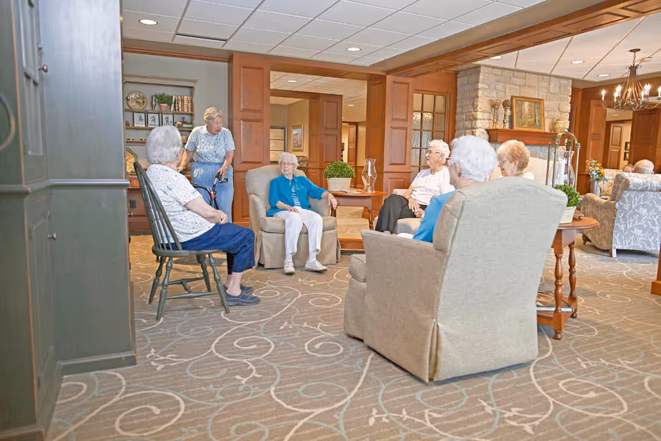 A group of elderly women sitting and conversing in a cozy living room area with armchairs and a wooden table. The room features a patterned carpet, wooden paneling, a stone fireplace, and decorative items on shelves and tables.